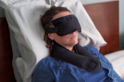 Person laying in a bed with an eye mask on and a black wrap around her neck 